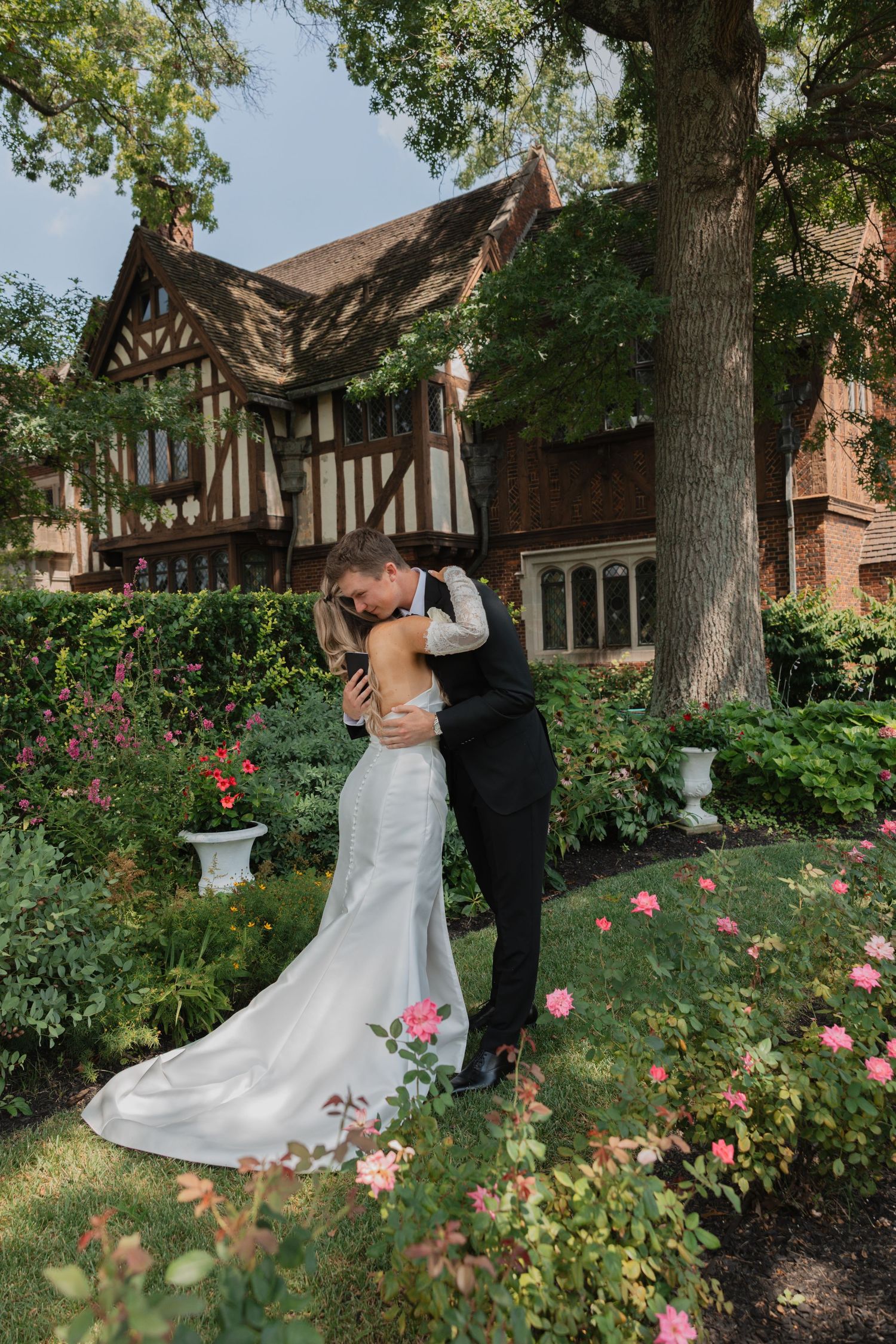 A romantic moment unfolds in a garden with blooming roses and Tudor-style architecture in the background.