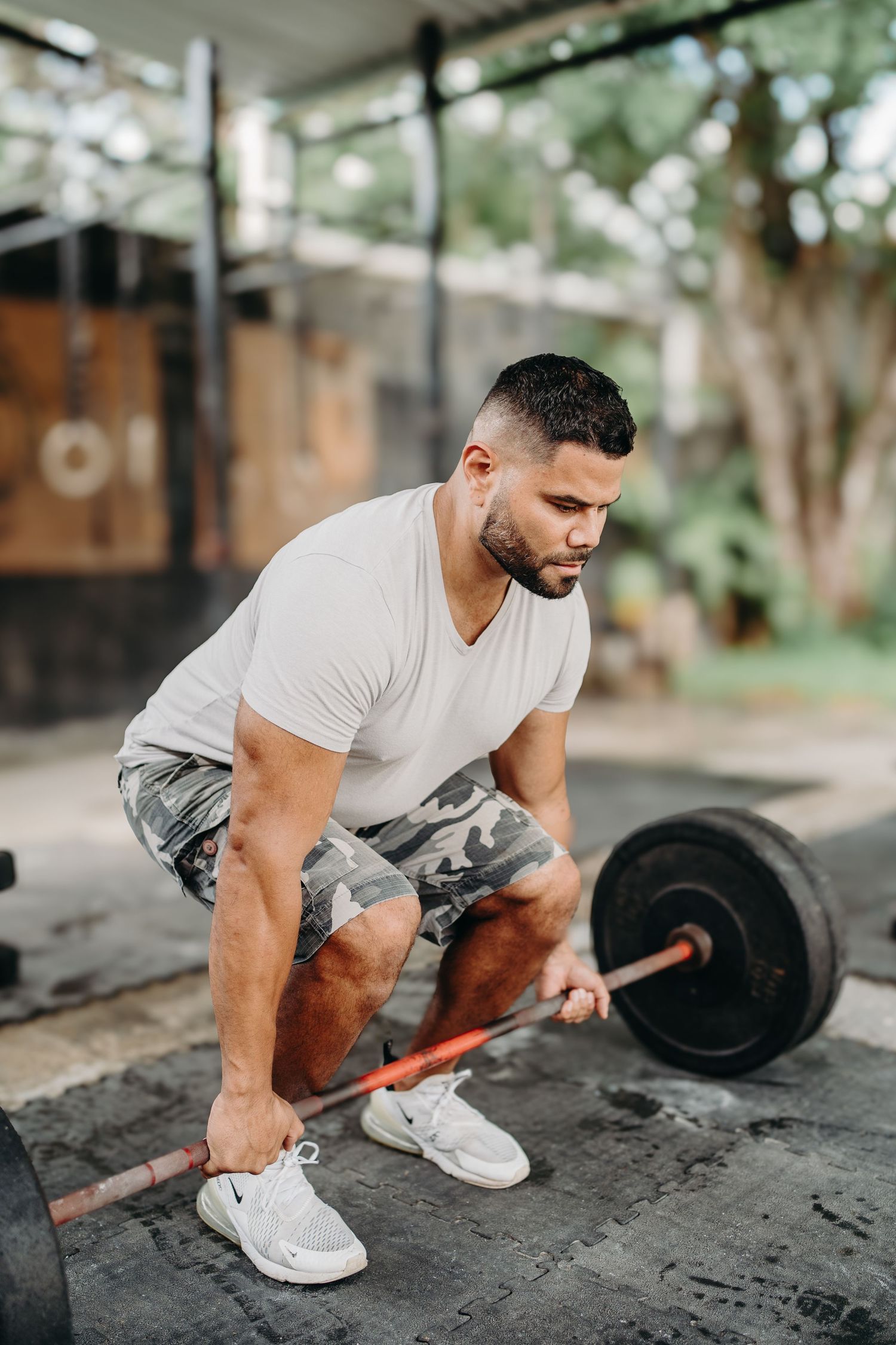 A person in a white v-neck shirt performs a barbell exercise sequence on a concrete floor outdoors.