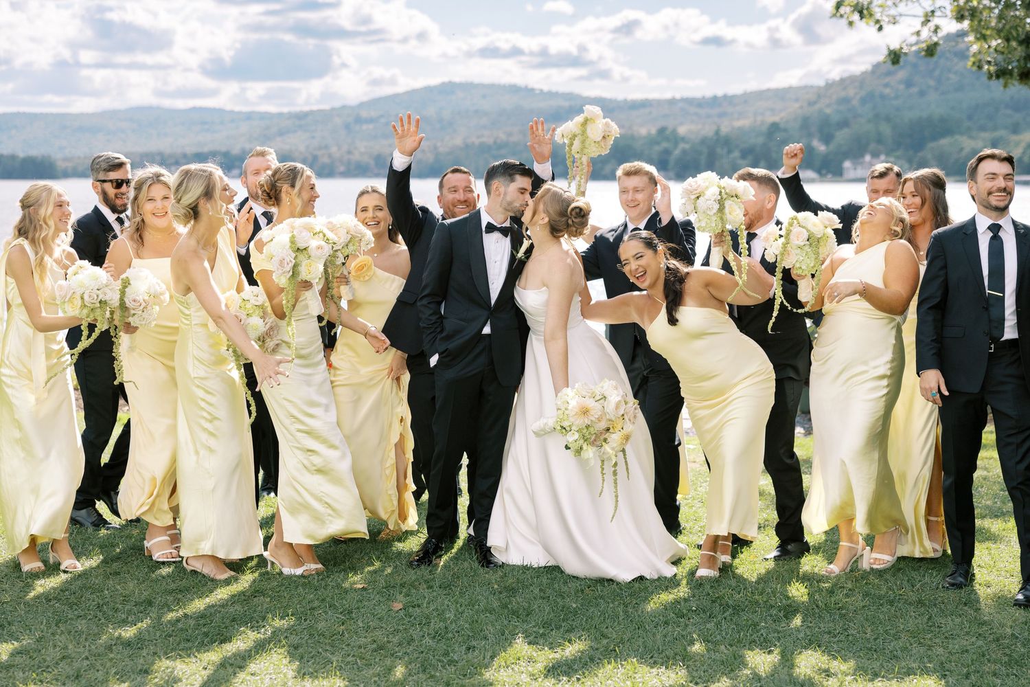 Wedding party celebrates outdoors with raised arms while standing in line formation against scenic mountain backdrop.