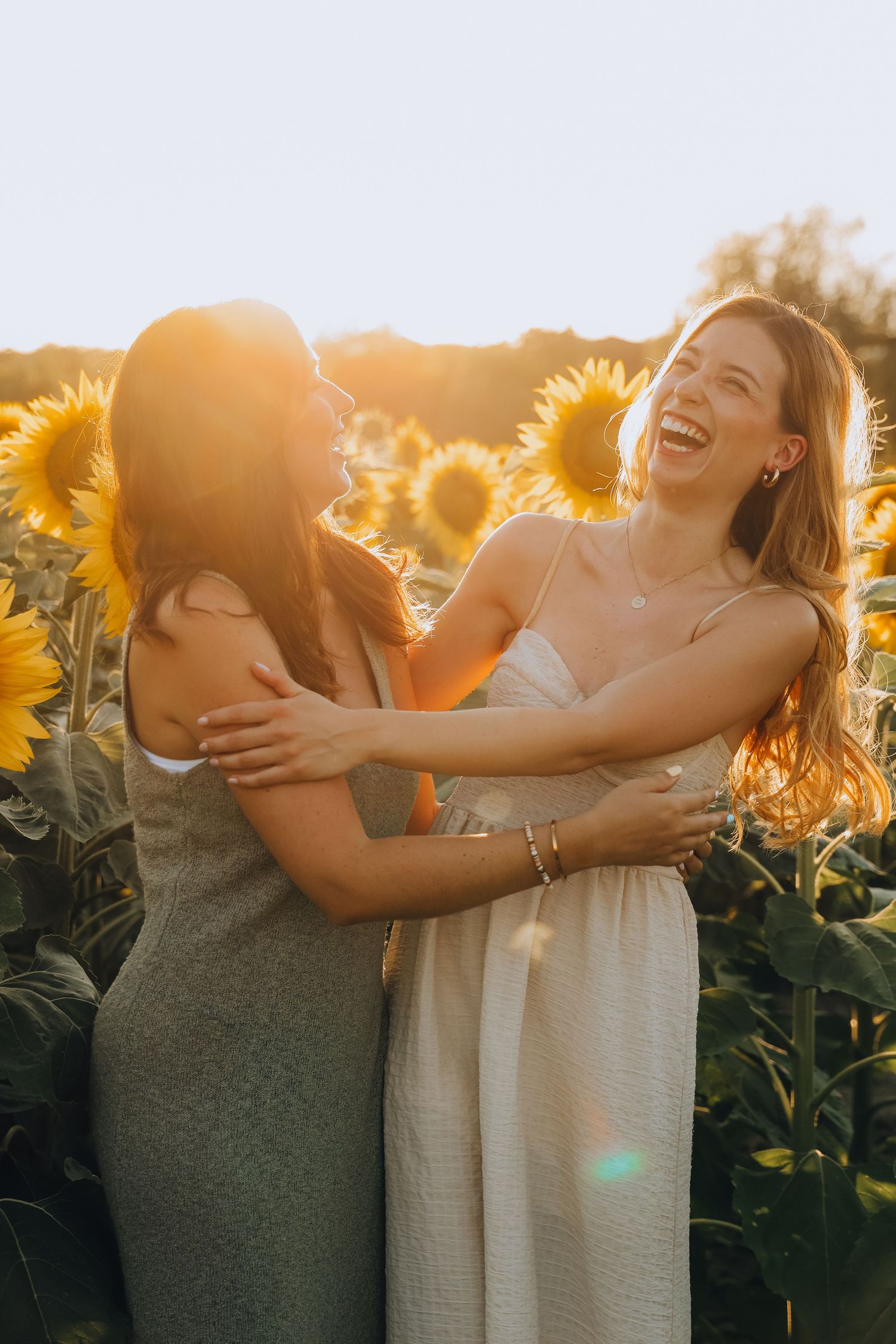Two women laughing and embracing in sunflower field with glowing sunset backlight.
