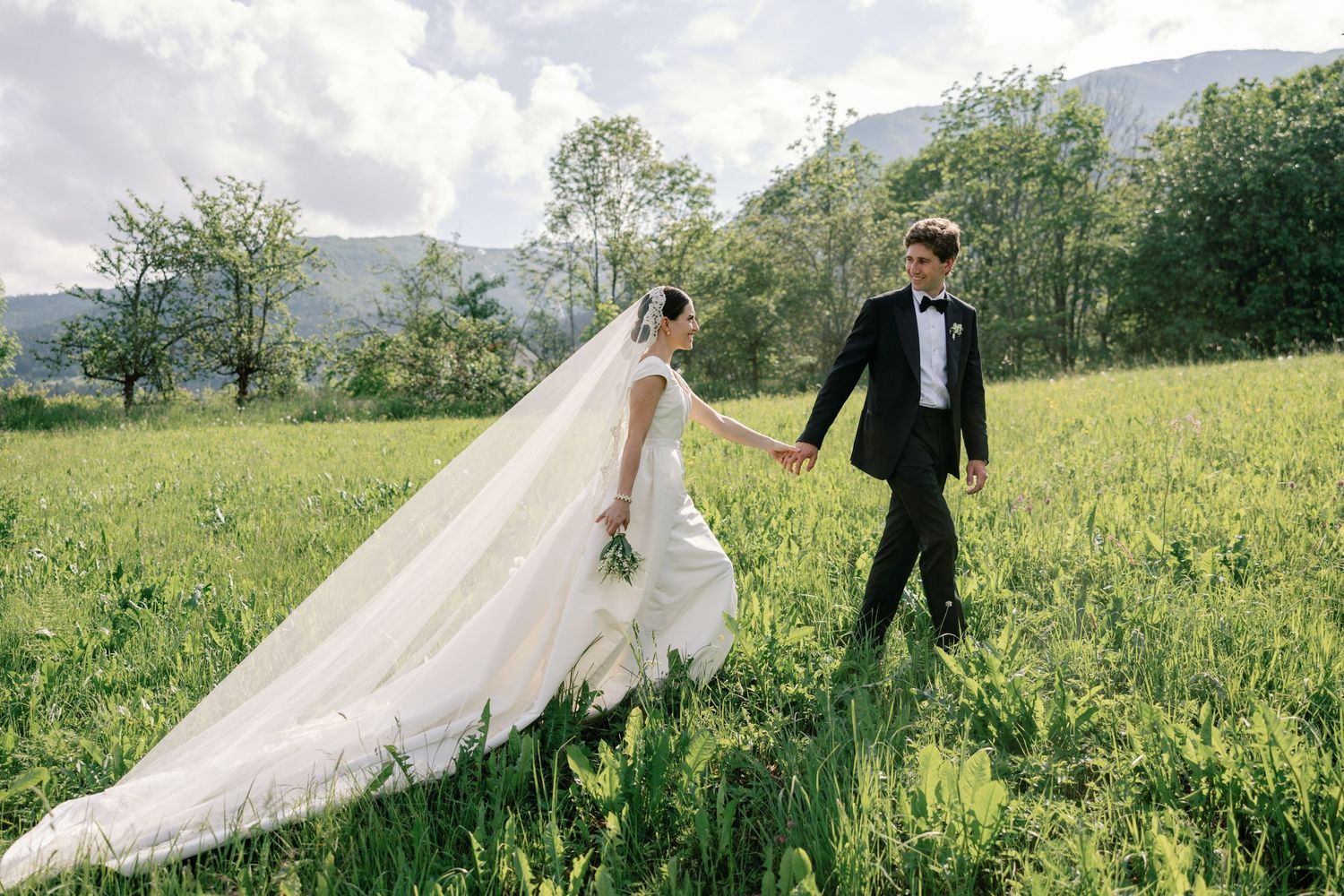 A romantic wedding couple strolls through a lush green meadow with mountains in the background.