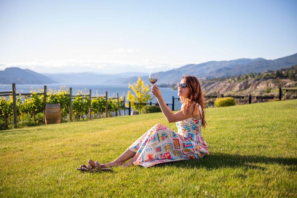 Woman in floral dress sitting on vineyard lawn, holding wine glass with mountain lake view backdrop on sunny day.