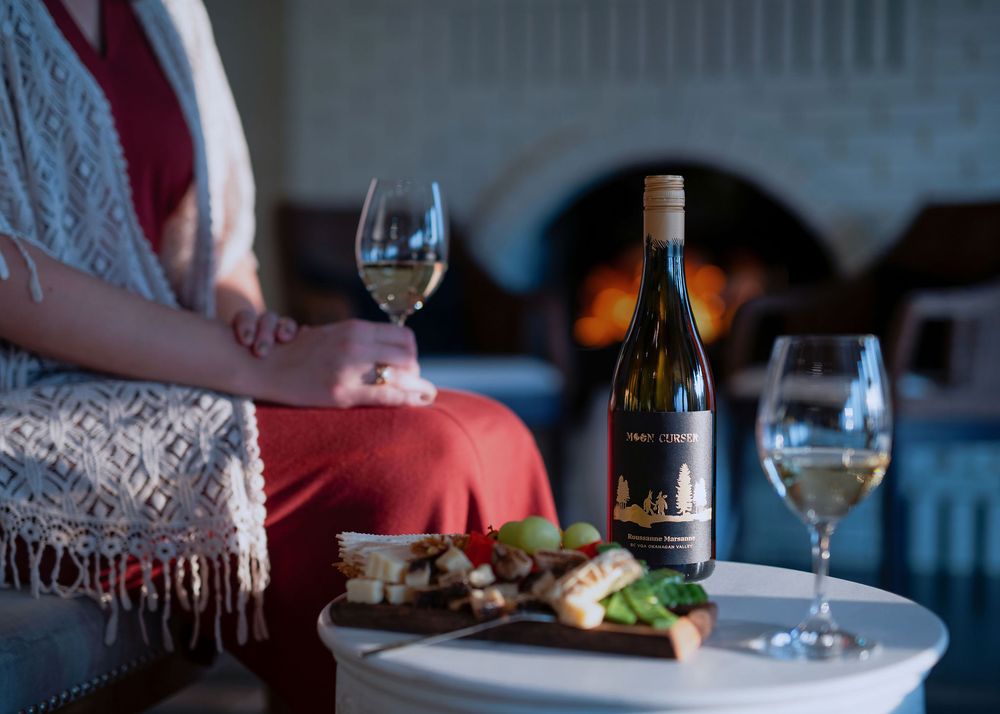 Woman in rust dress with lace shawl holding white wine by fireplace with Moon Curser bottle and charcuterie - Okanagan brand photo.