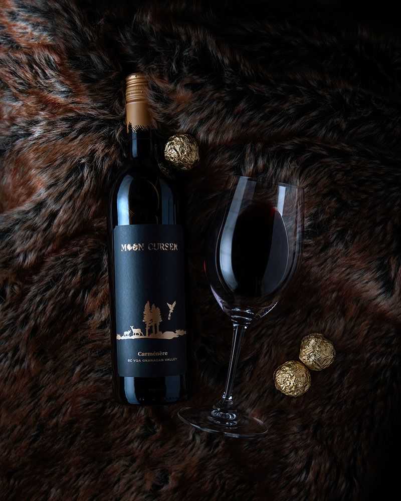 Top-down flat lay of Moon Curser wine bottle, tipped glass with red wine, and gold chocolates on brown fur - Okanagan brand photo.