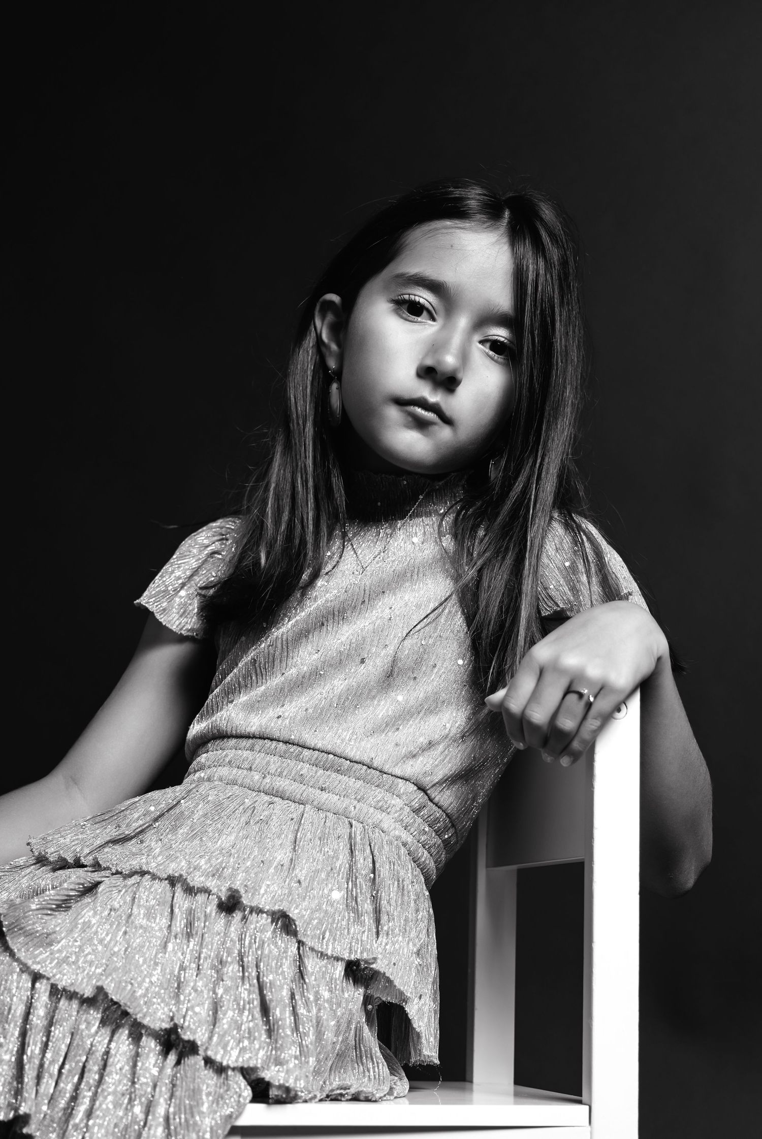 Black and white portrait of someone wearing a light-colored ruffled dress while posing against a dark background.