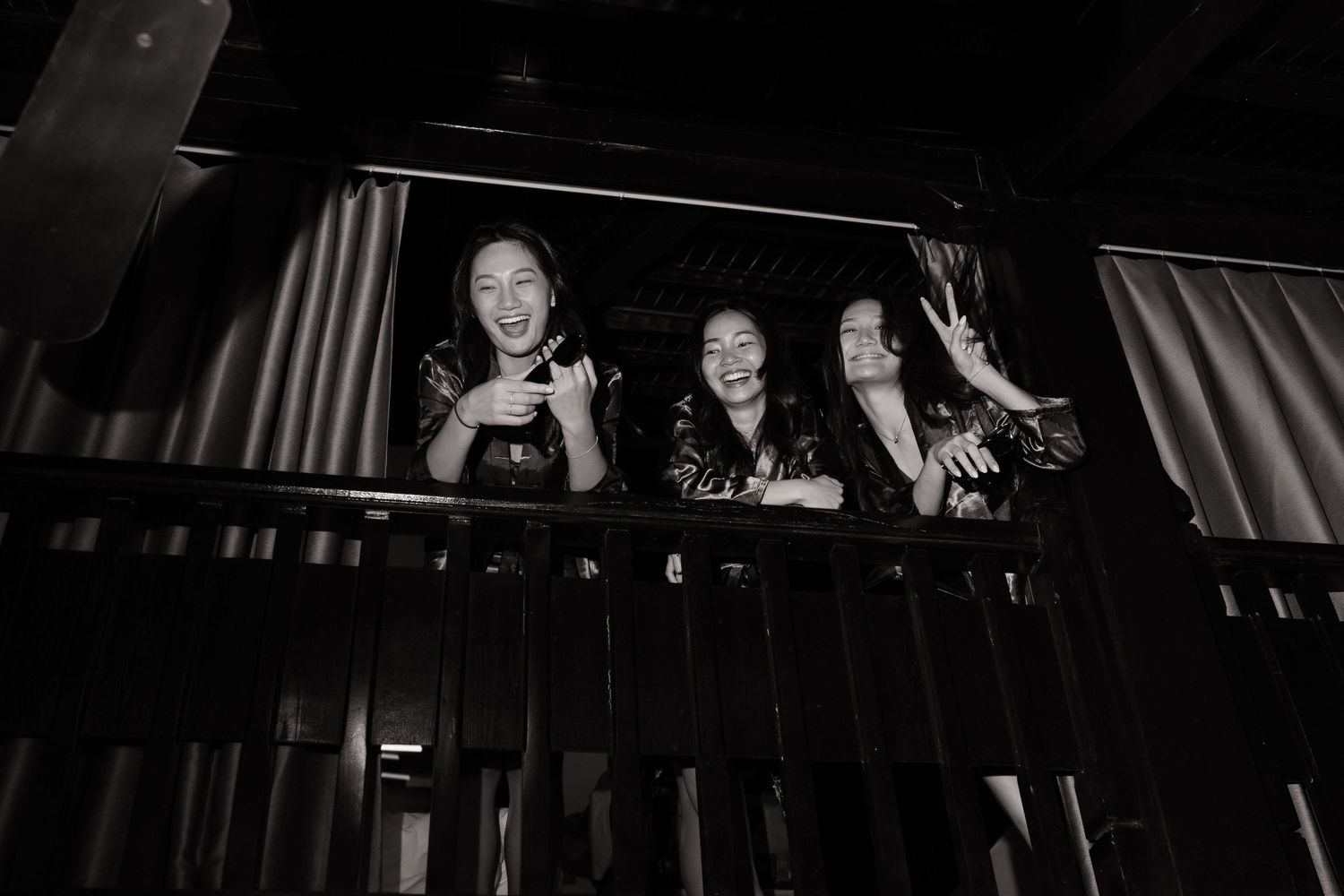 Three women in robes laughing and posing on a dark balcony railing in black and white.