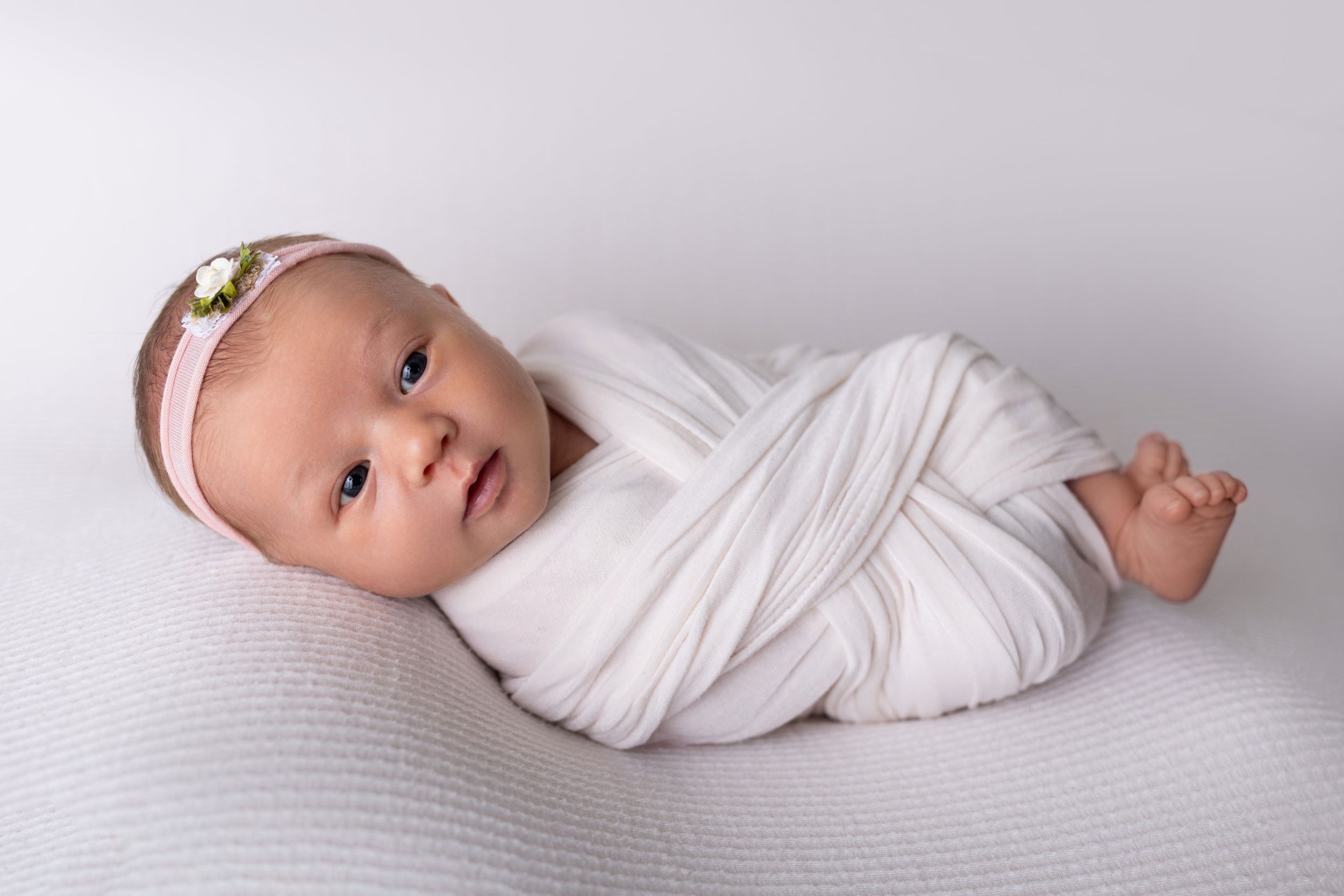 A newborn baby lies on a soft gray blanket wearing a white wrap in a minimalist photography style.