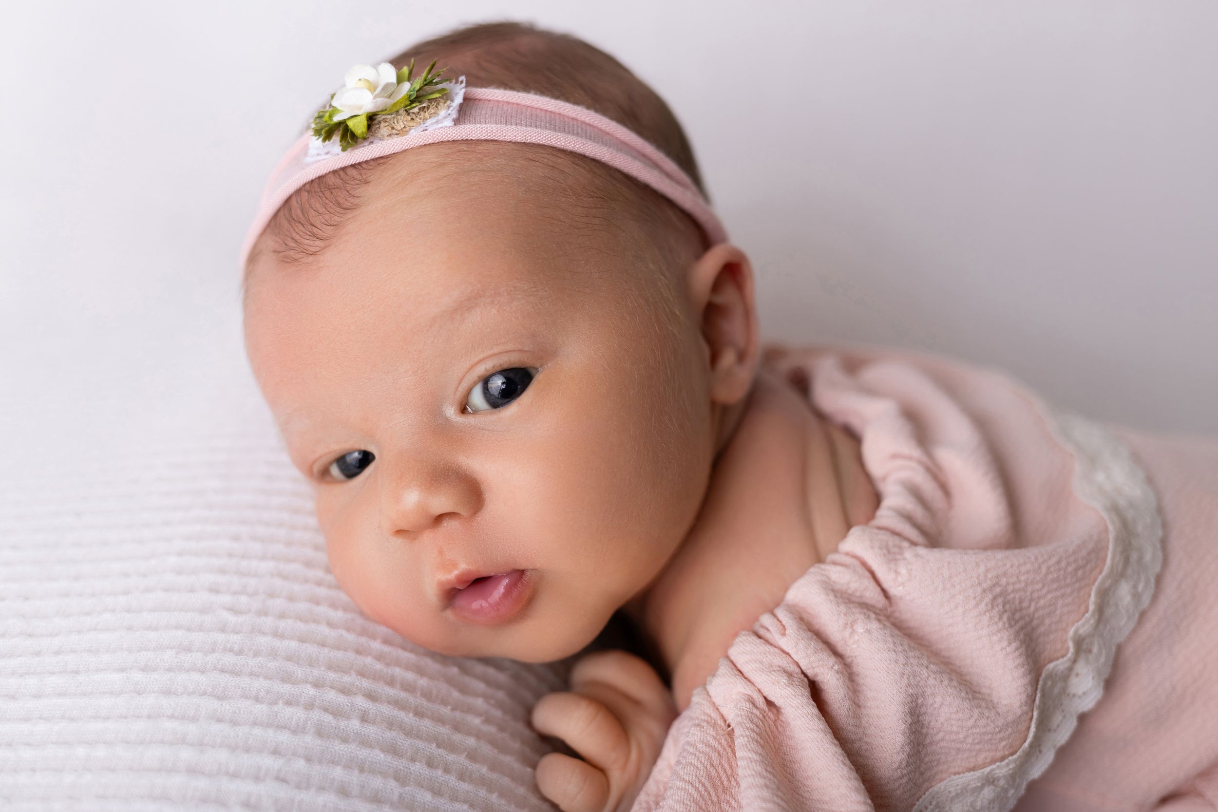 Newborn baby wearing pink headband lying on soft white blanket while being photographed during professional portrait session.