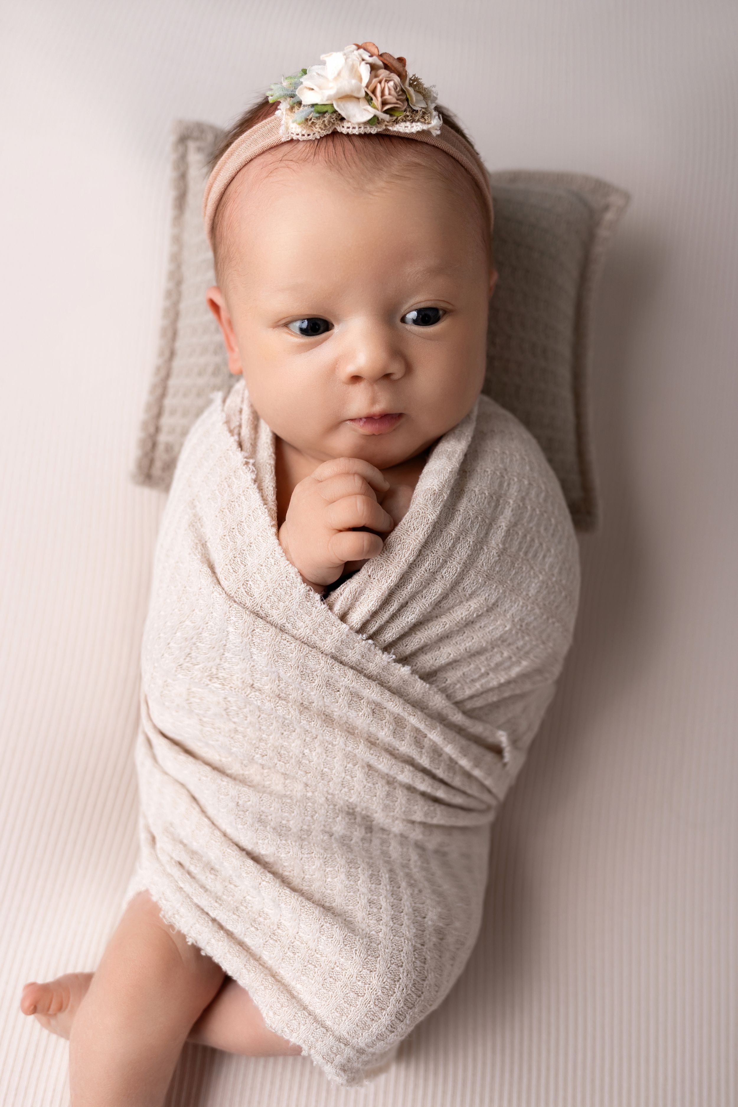 Newborn wrapped in beige muslin fabric lying on white backdrop wearing a delicate floral headband.