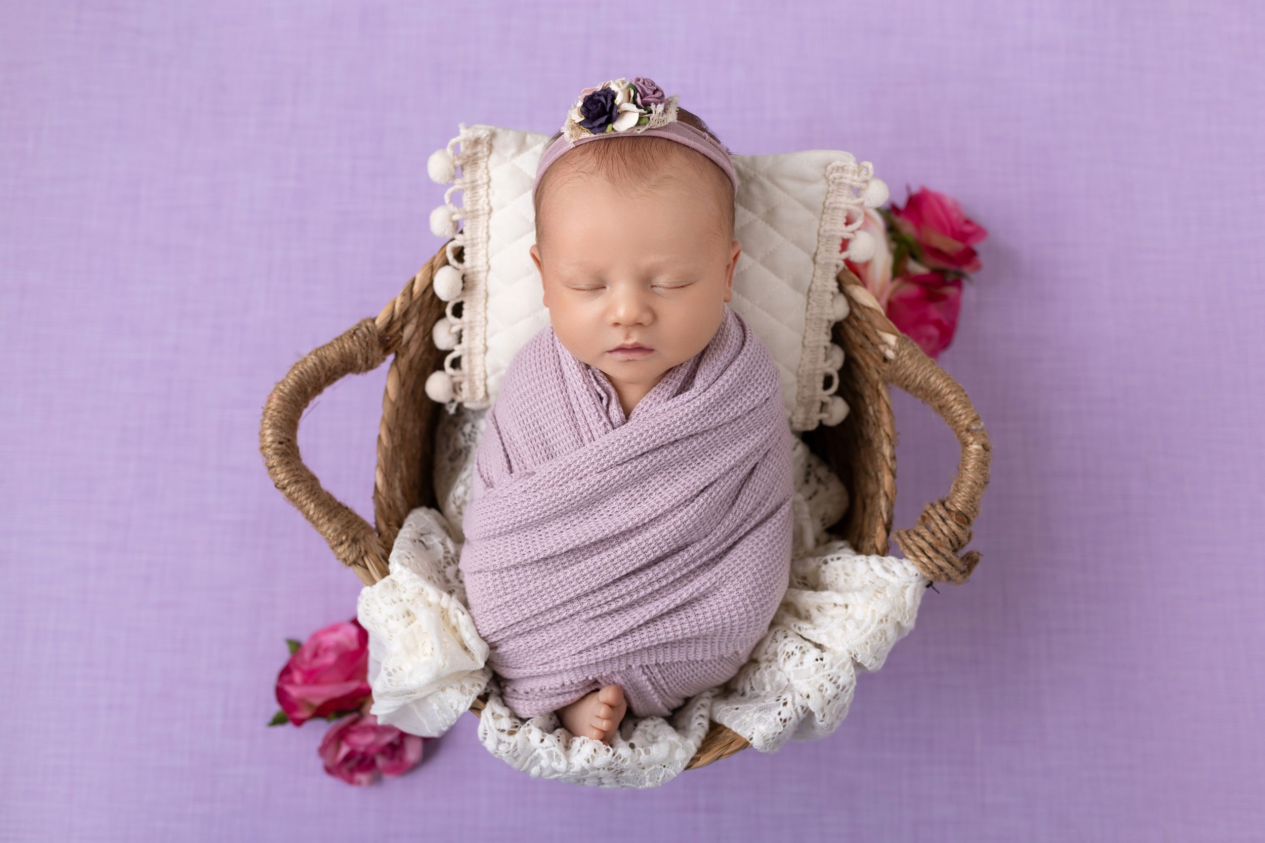 Sleeping newborn wrapped in lavender fabric nestled in woven basket with pink flower accents.