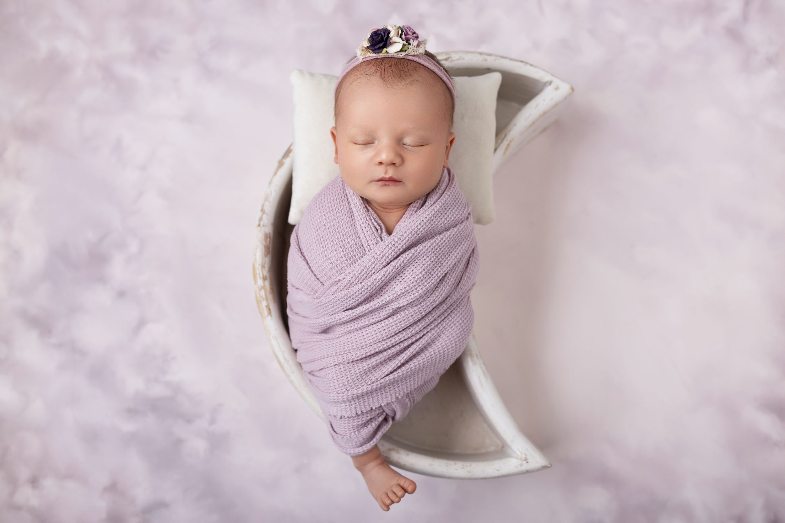 Baby in lavender swaddle wrap posed on textured backdrop during professional newborn photoshoot.