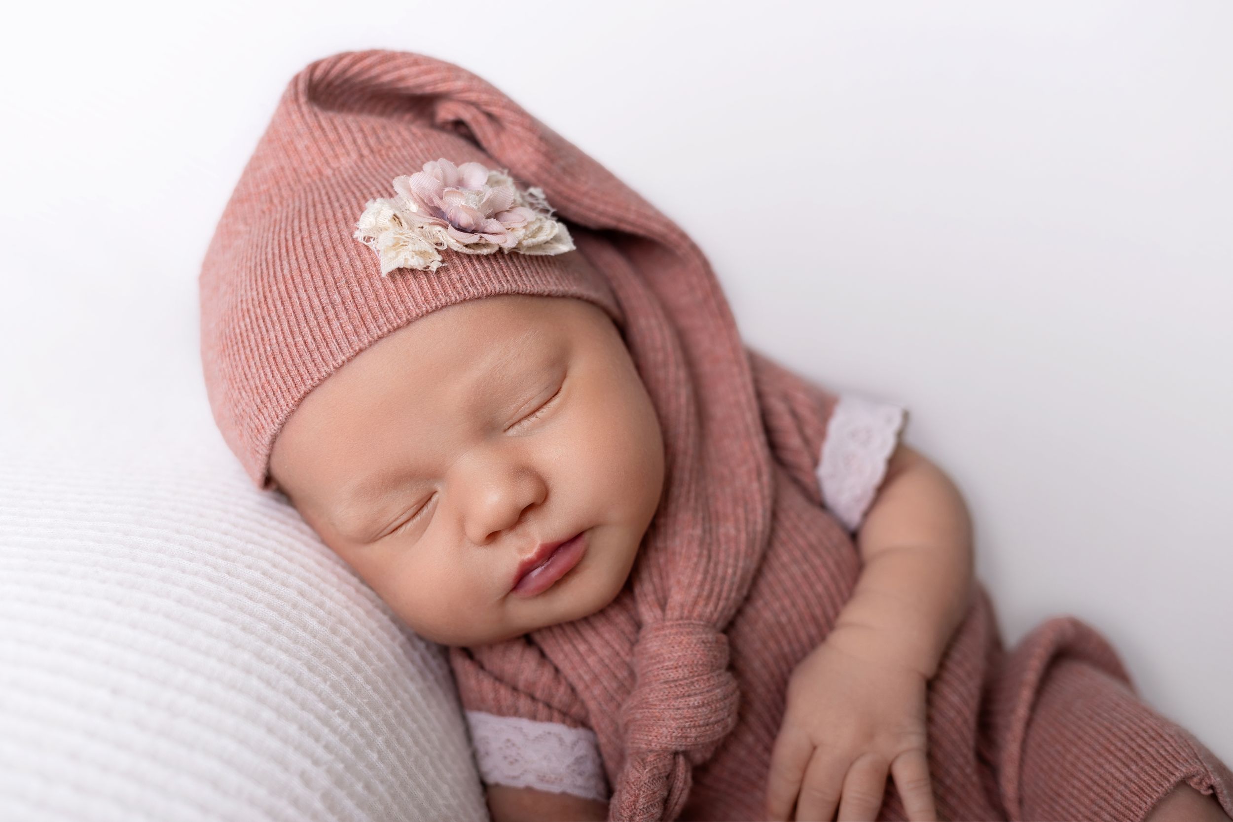Peaceful sleeping newborn wearing dusty rose knit hat and wrap against white textured background.