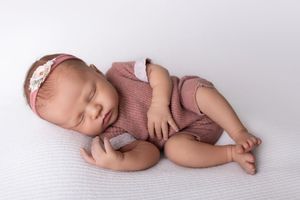 Sleeping newborn baby in mauve knitted outfit lying on white backdrop.