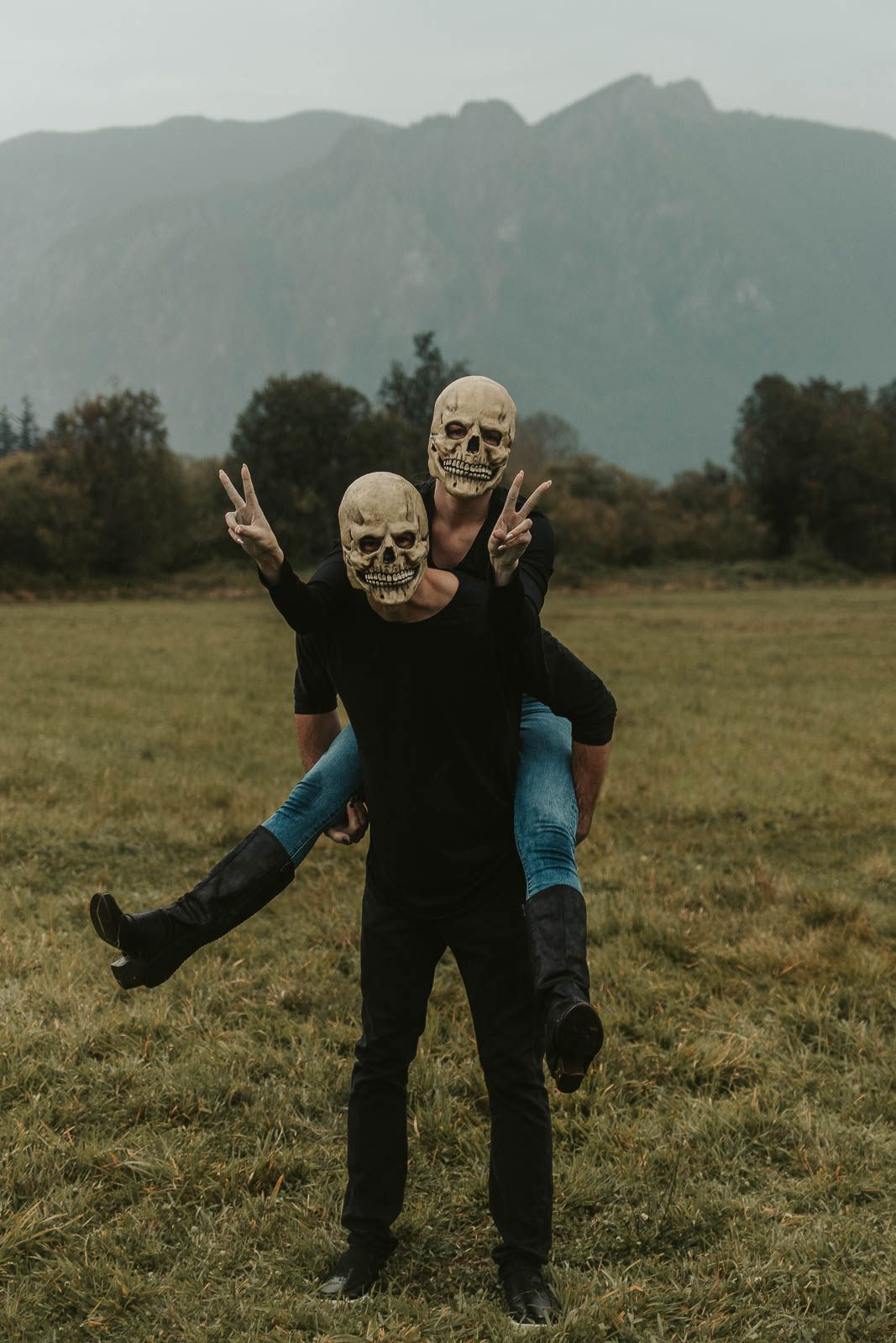 spooky Halloween couples photo shoot man holding woman in a piggy back ride both wearing skull mask in a foggy field and mountain background