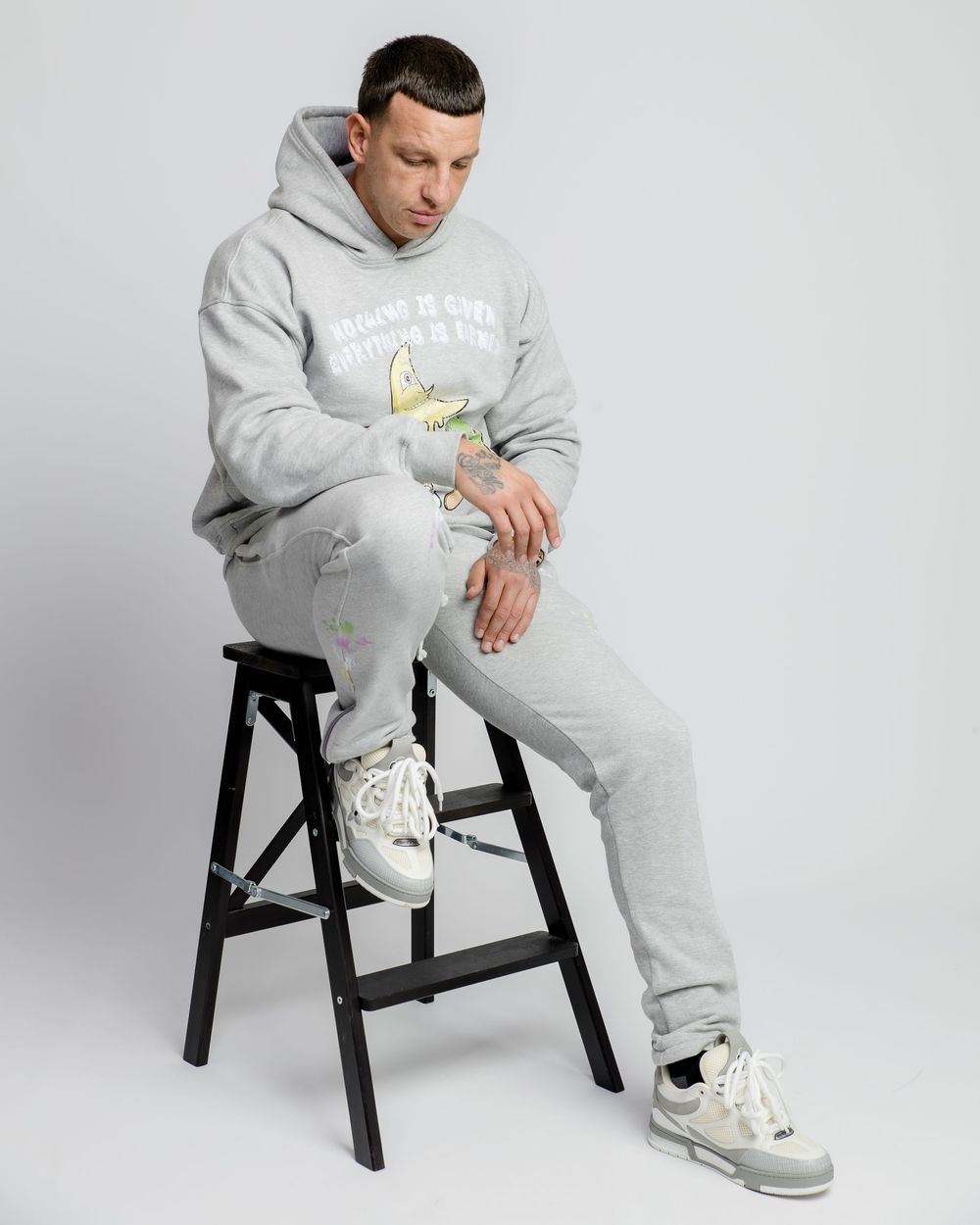 Person in gray sweatsuit and white sneakers sitting on black stool against white backdrop.