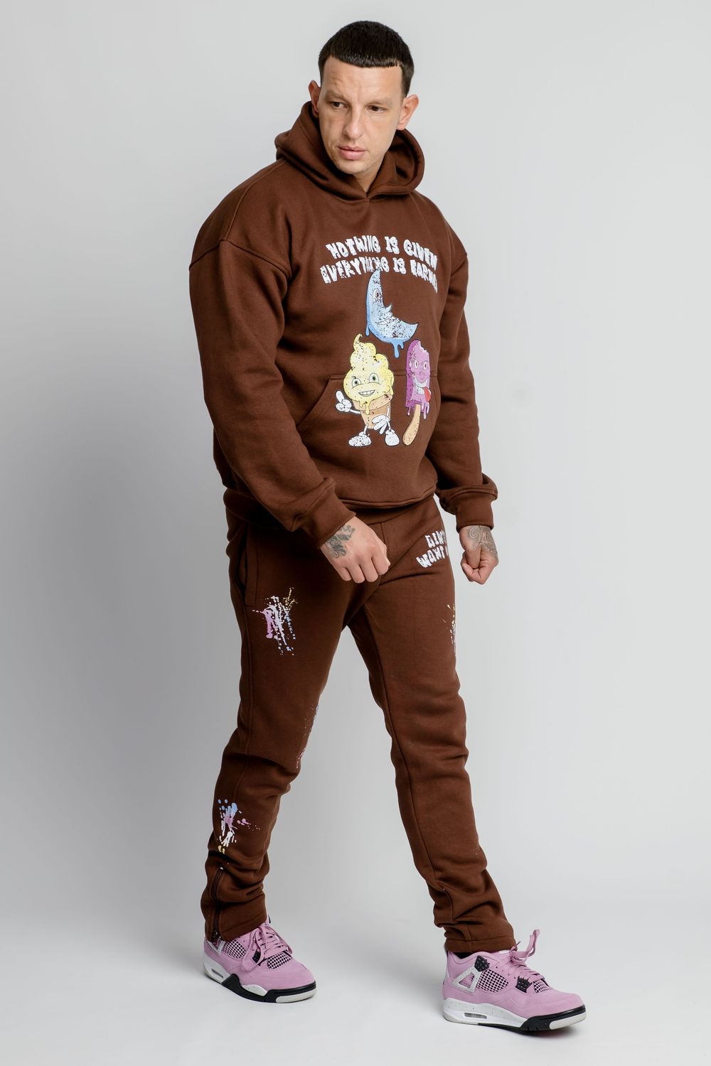 Model showcases brown sweatsuit with cartoon character graphic and purple sneakers in studio setting.