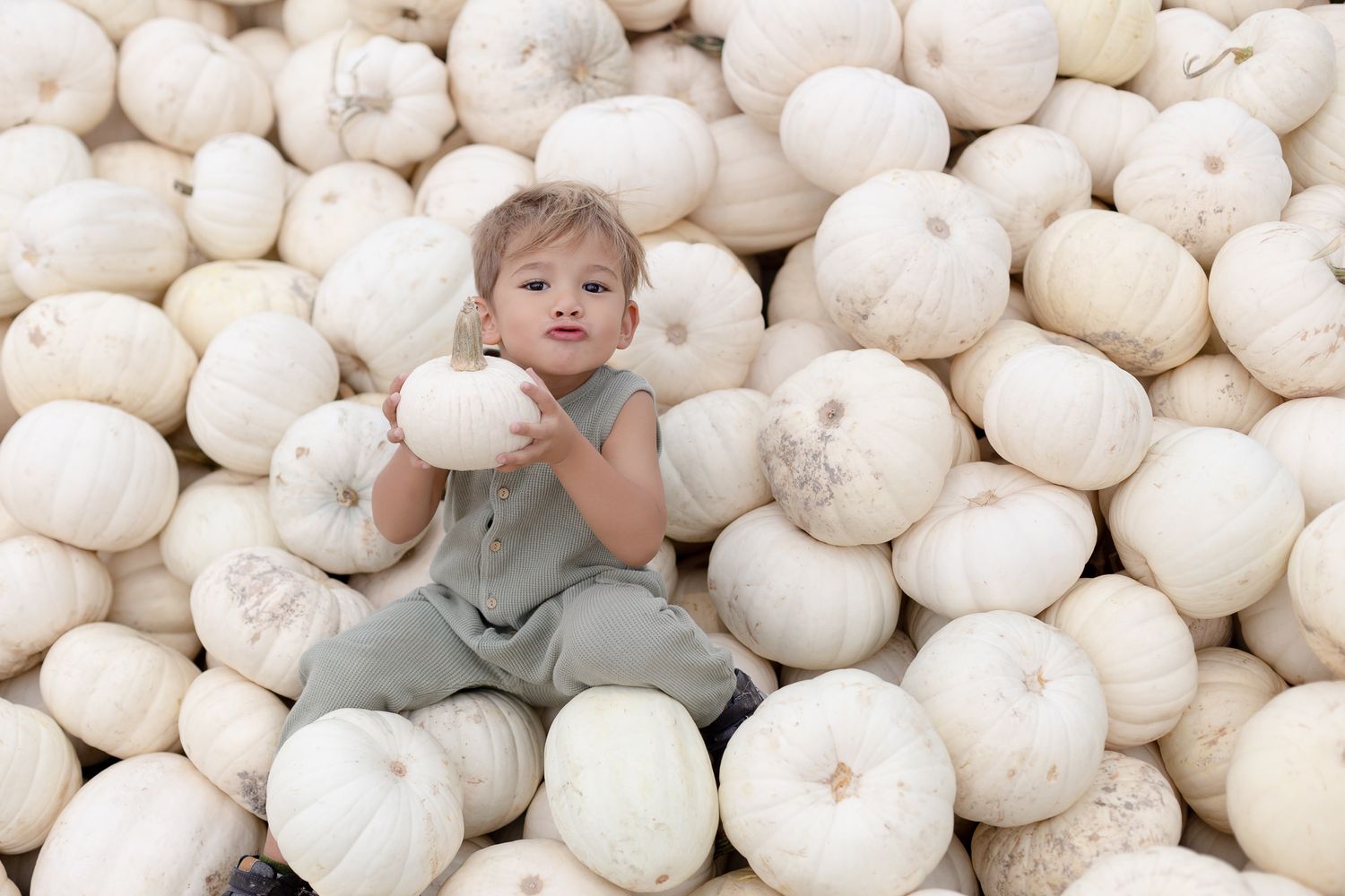 Small child in gray overalls sits among large white pumpkins at a pumpkin patch.