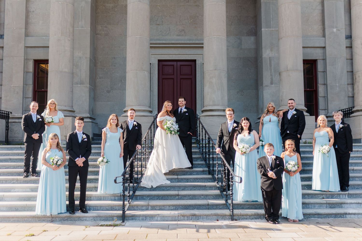 Wedding party in light blue bridesmaid dresses and formal attire poses on grand stone steps of classical building.