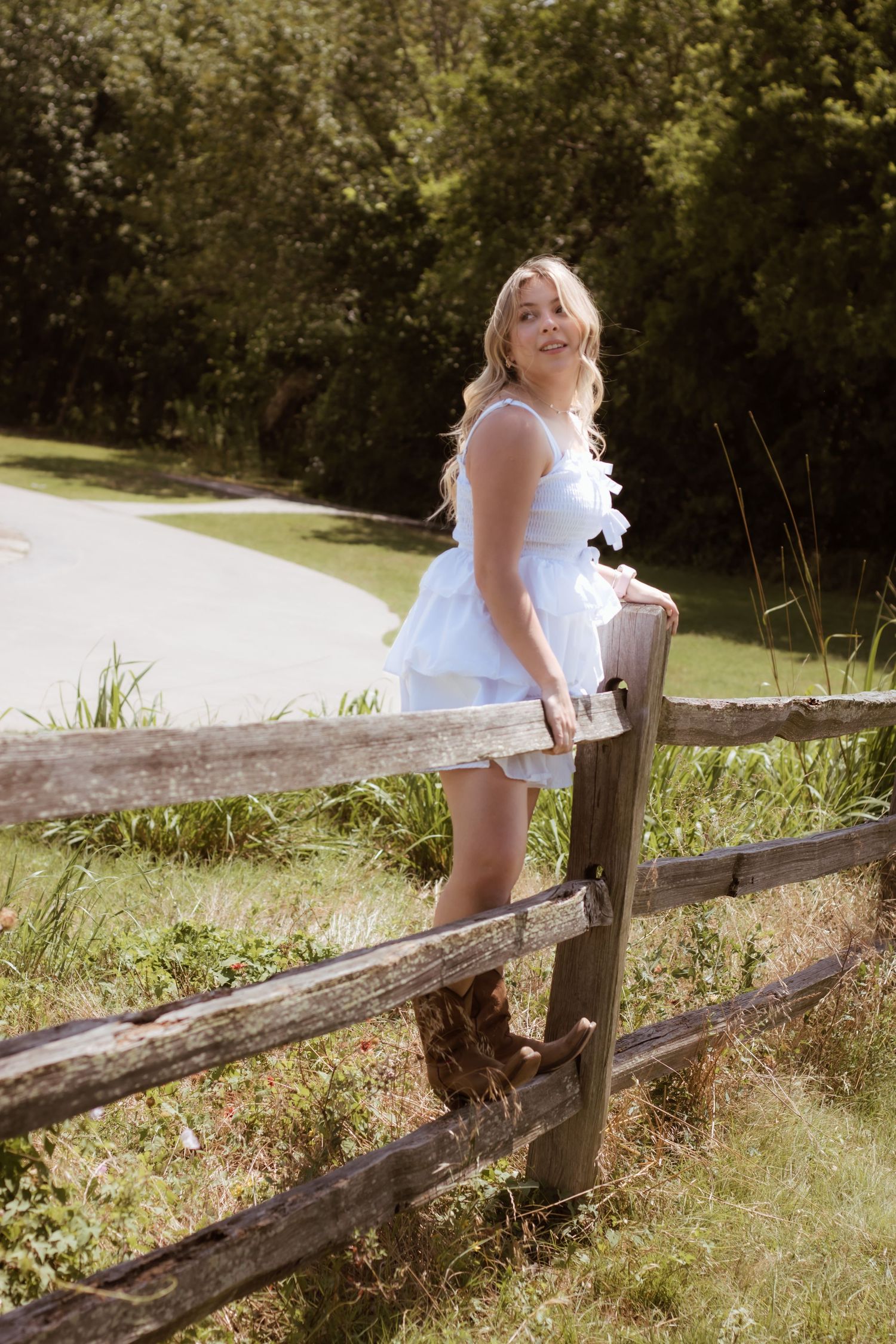 A person in a white summer dress and boots leans on a rustic wooden fence near a pond on a sunny day.