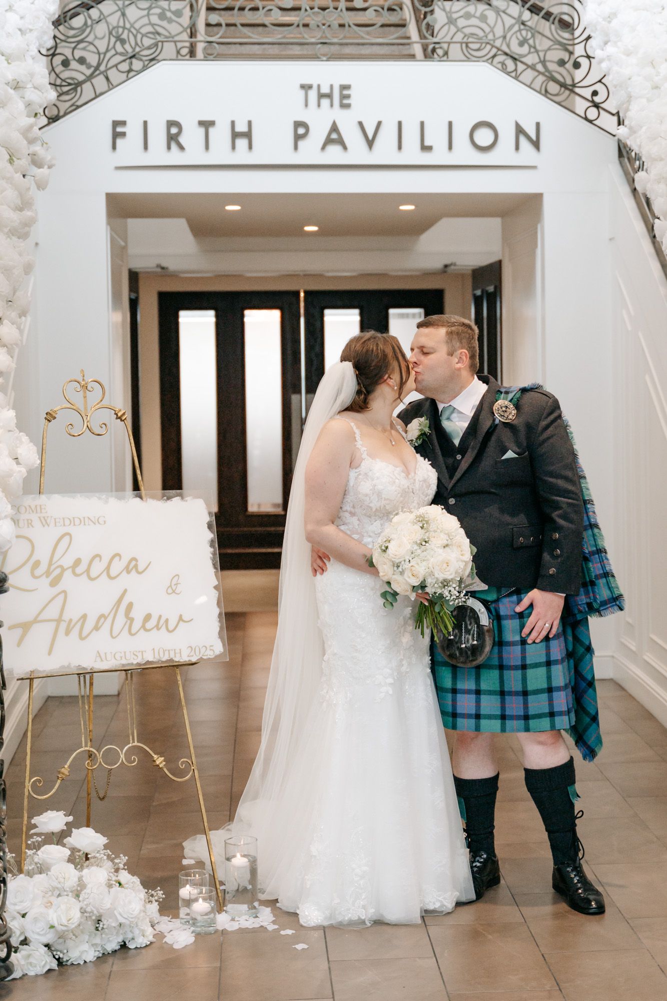 A couple poses in front of the Firth Pavilion sign with the groom wearing a traditional Scottish kilt.