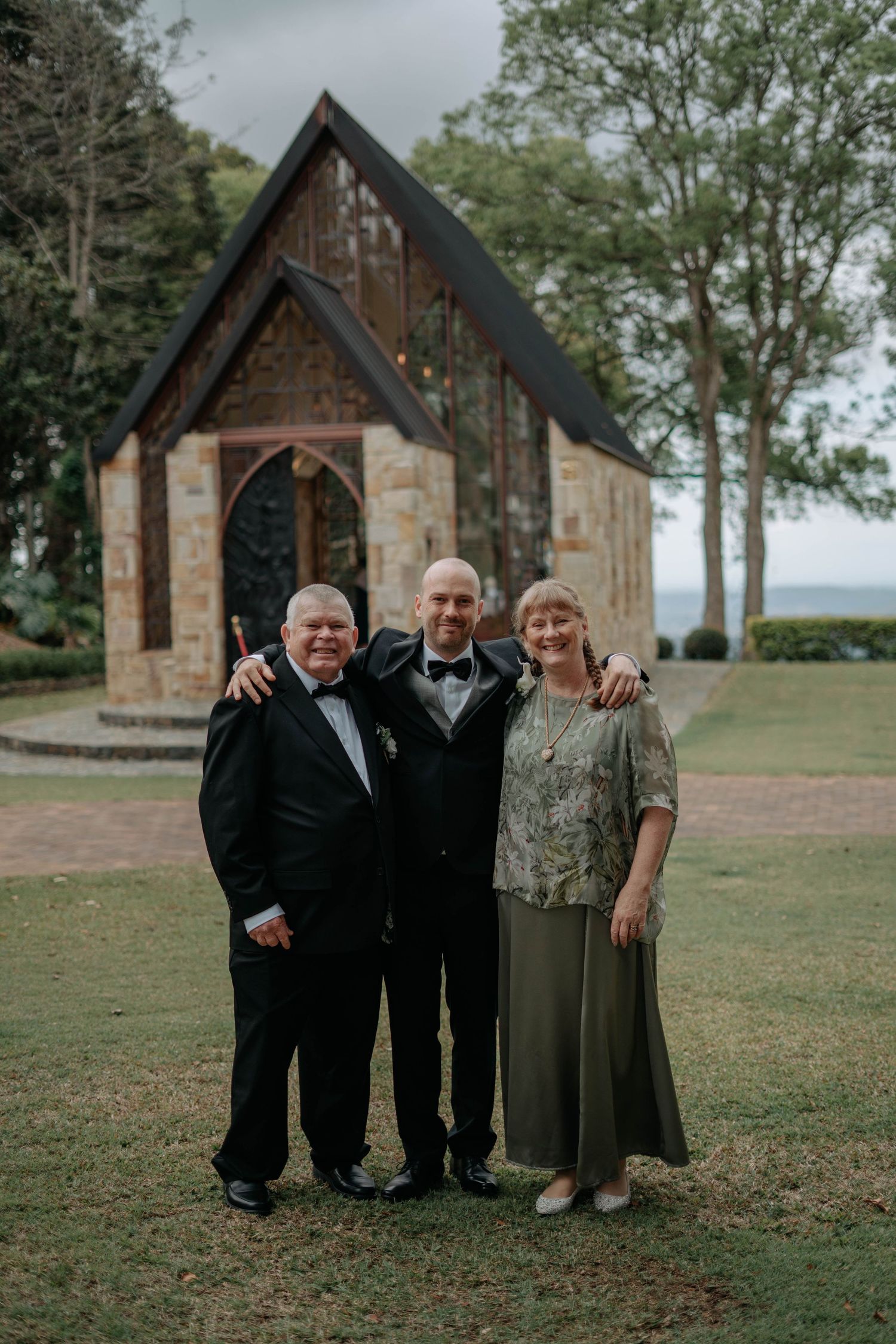 Three people in formal attire pose together in front of a charming stone chapel with a dramatic peaked roof on a cloudy day.