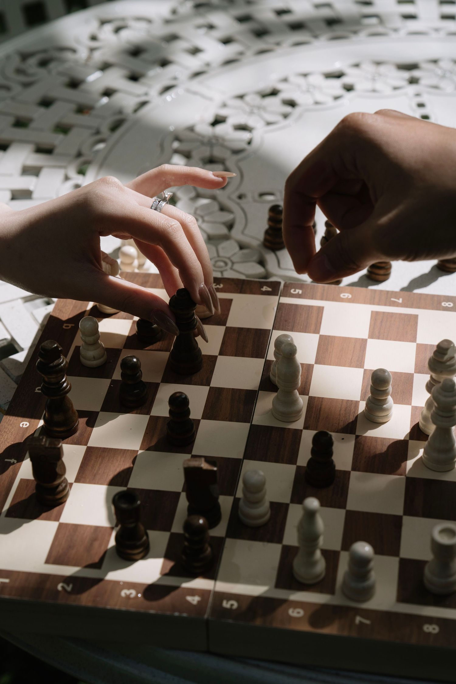 Two people playing chess on a wooden board, with hands reaching to move pieces in dramatic lighting.