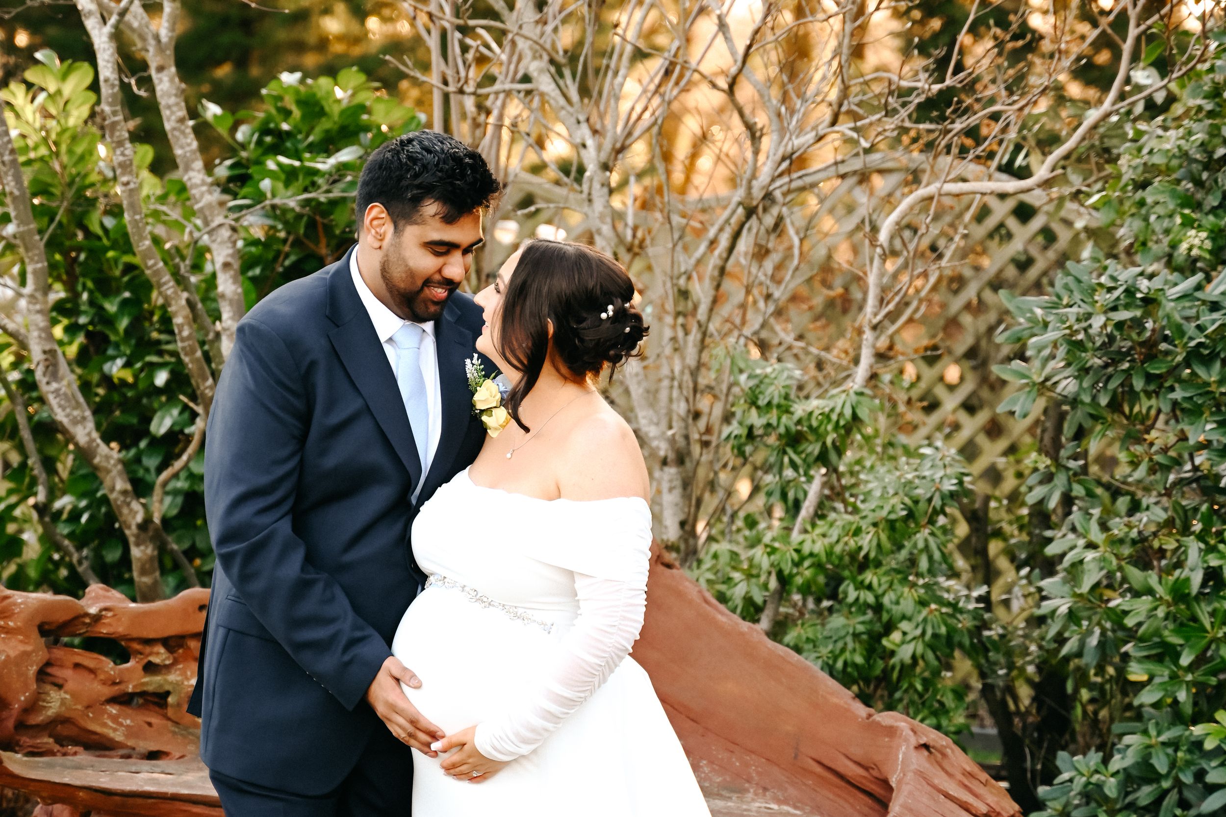 Pregnant bride in white off-shoulder gown with groom in navy suit sharing tender moment outdoors among lush greenery.
