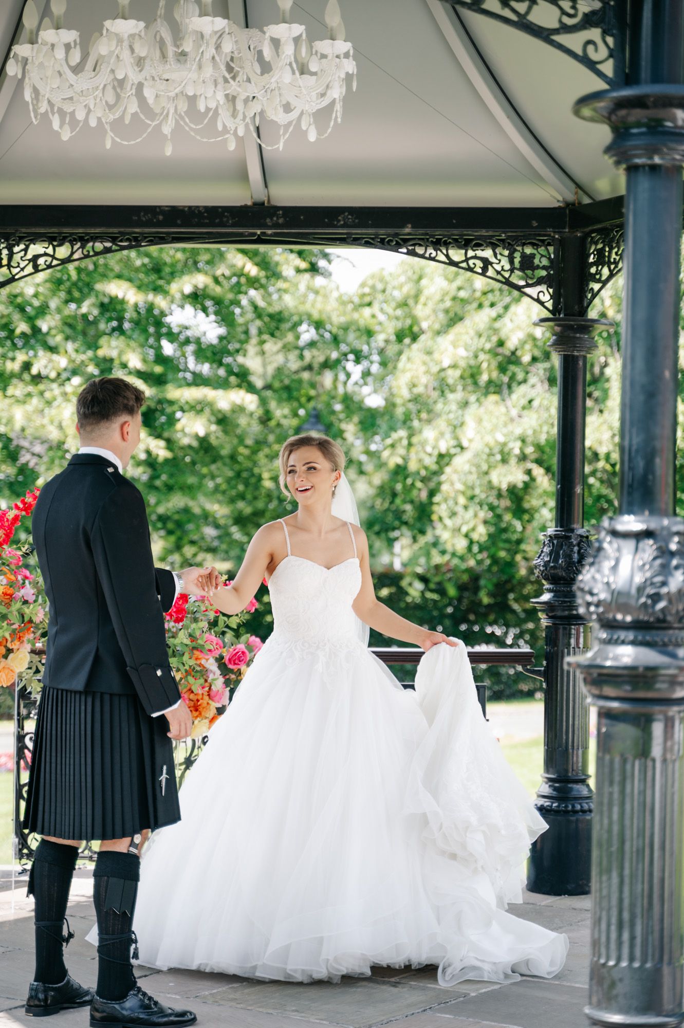 A wedding couple shares intimate moments in a white gazebo decorated with floral arrangements and chandeliers.