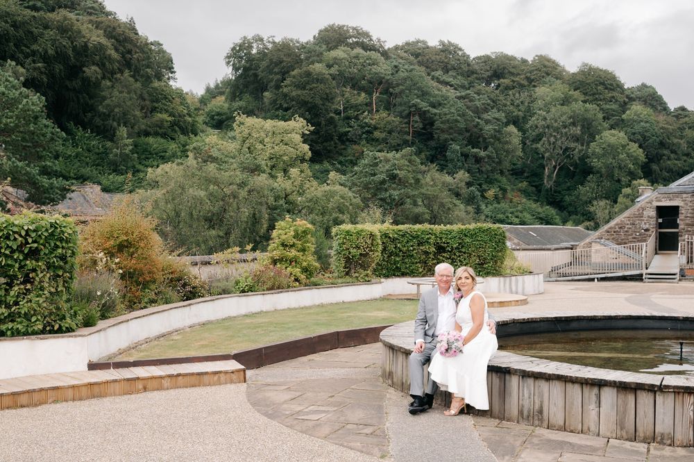 Couple sitting together by a fountain in a lush garden setting on a cloudy day.