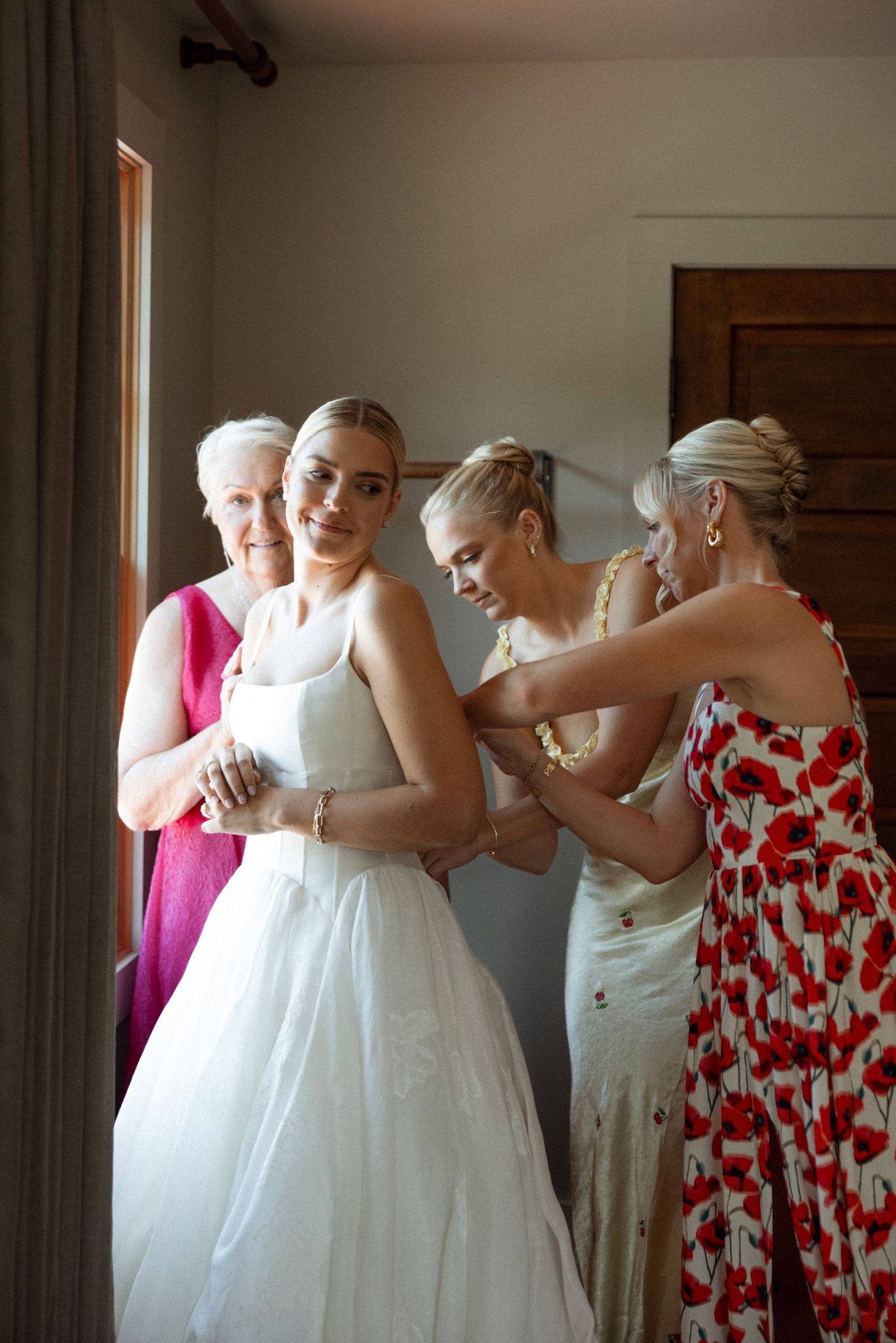 People assist with preparations in a bridal suite, helping with a white wedding dress.