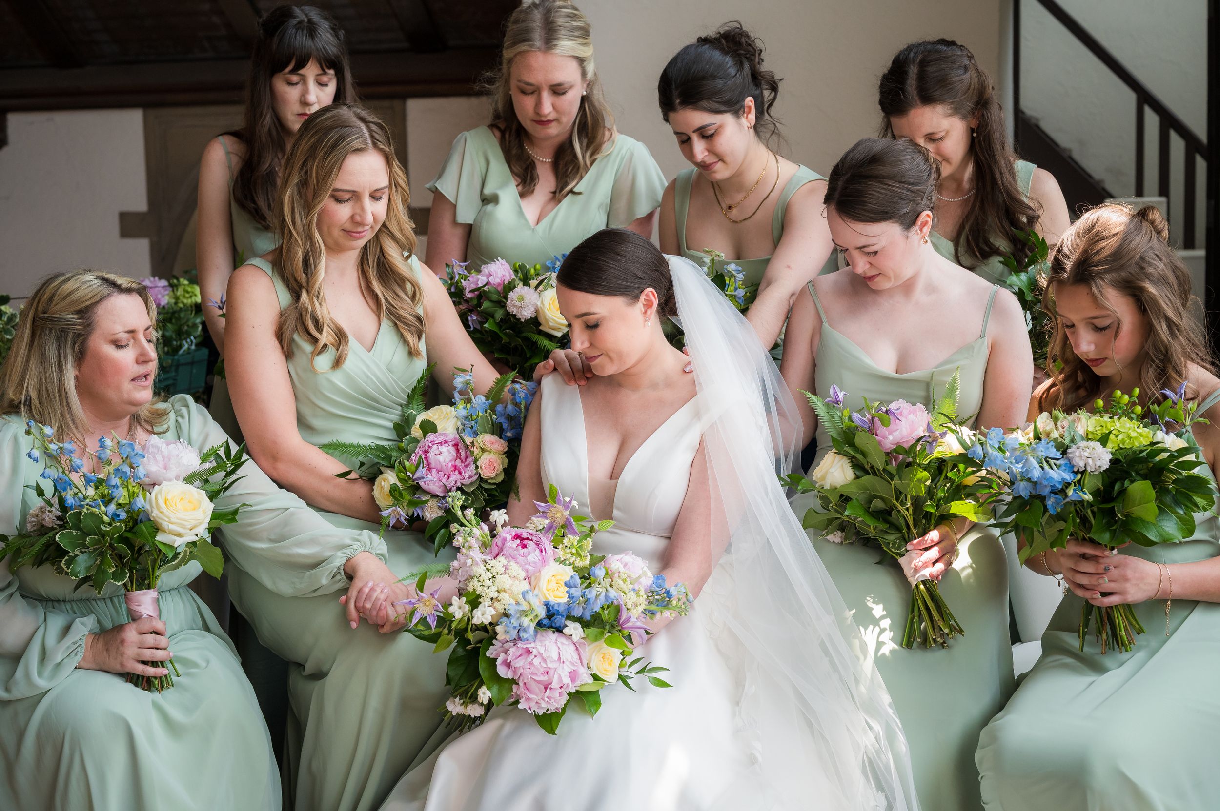 Bridesmaids in sage green dresses pose together holding colorful bouquets of pink and purple flowers.