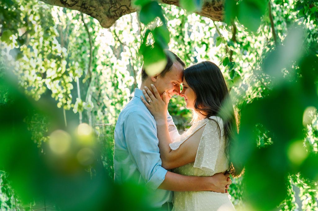 Two people share a romantic kiss in a sunlit garden setting with green leaves framing the intimate moment.