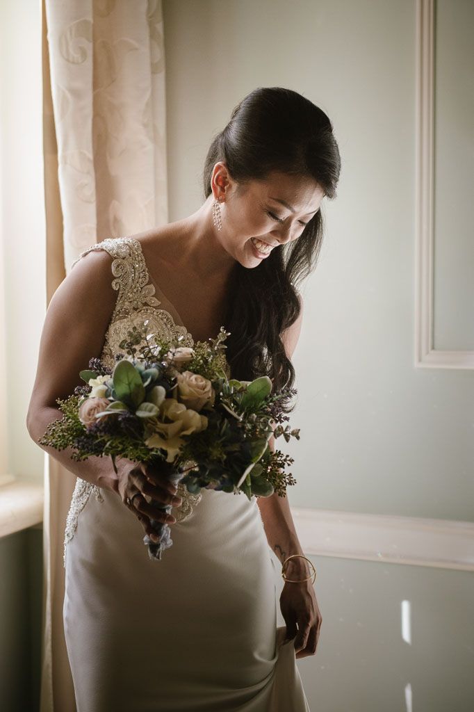 A bride in a white wedding dress holds a soft-toned floral bouquet in natural window light.