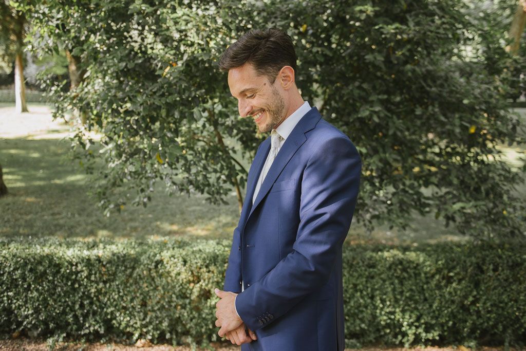 A row of groomsmen in navy suits stand together outdoors against a green garden backdrop.