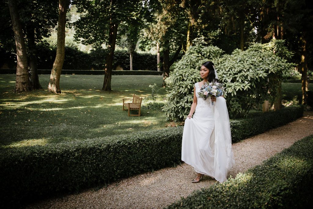 A wedding couple poses together in a lush garden setting surrounded by blooming white floral trees and gravel pathways.
