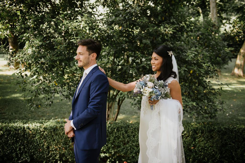 A bride in a white dress holding a bouquet stands in a garden with someone wearing a navy blue suit.