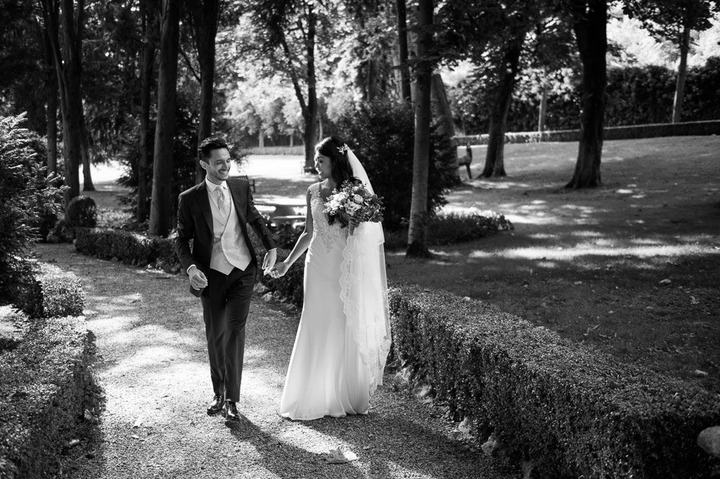 Couple holding hands walking through a lush garden path with trees and natural sunlight filtering through.