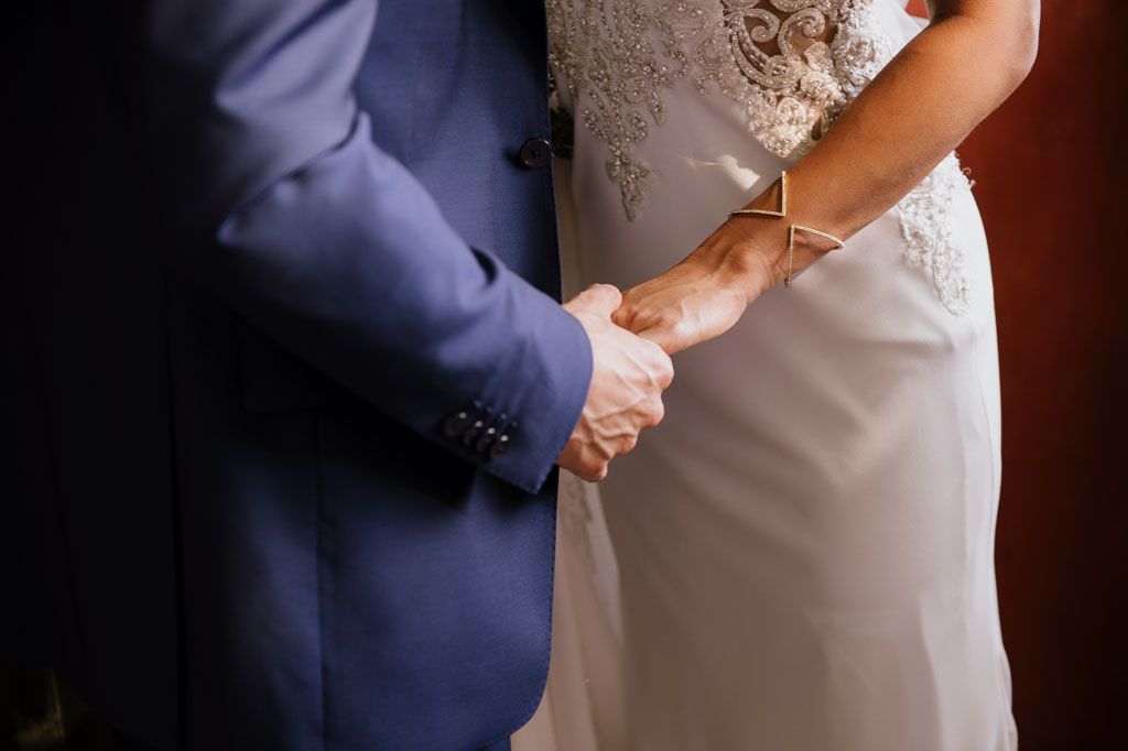 A couple's hands clasped together during an outdoor wedding ceremony, with bride in a white gown and groom in navy suit.