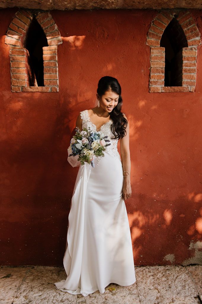 Woman in white wedding dress poses against rustic terracotta wall with small arch windows in Mexican hacienda setting.