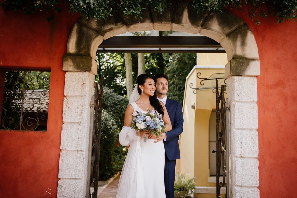 A couple in wedding attire stands under a rustic stone archway against a vibrant red wall during their ceremony.