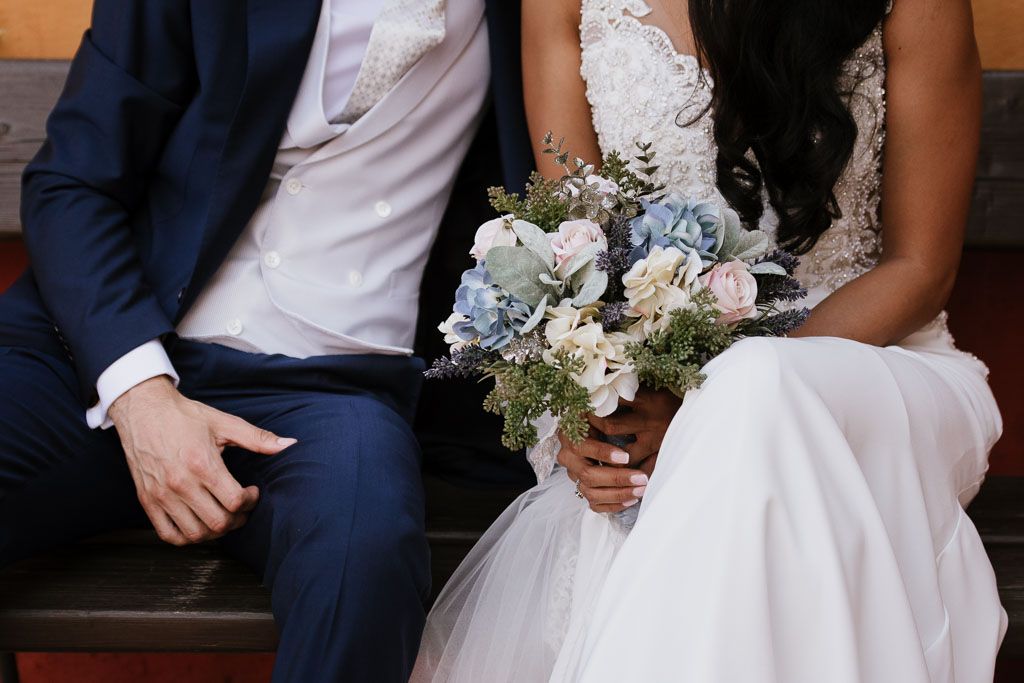A couple sits together on wooden steps as the bride holds a soft blue and white floral wedding bouquet.