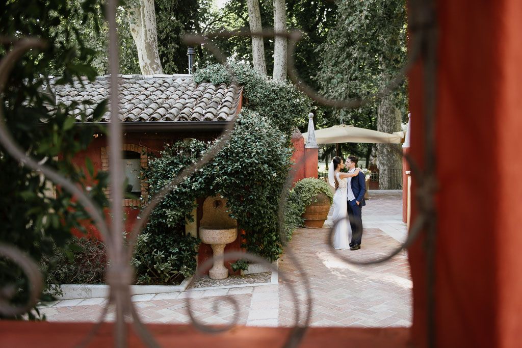 Wedding couple strolls through scenic garden pathway lined with red pillars and lush greenery.