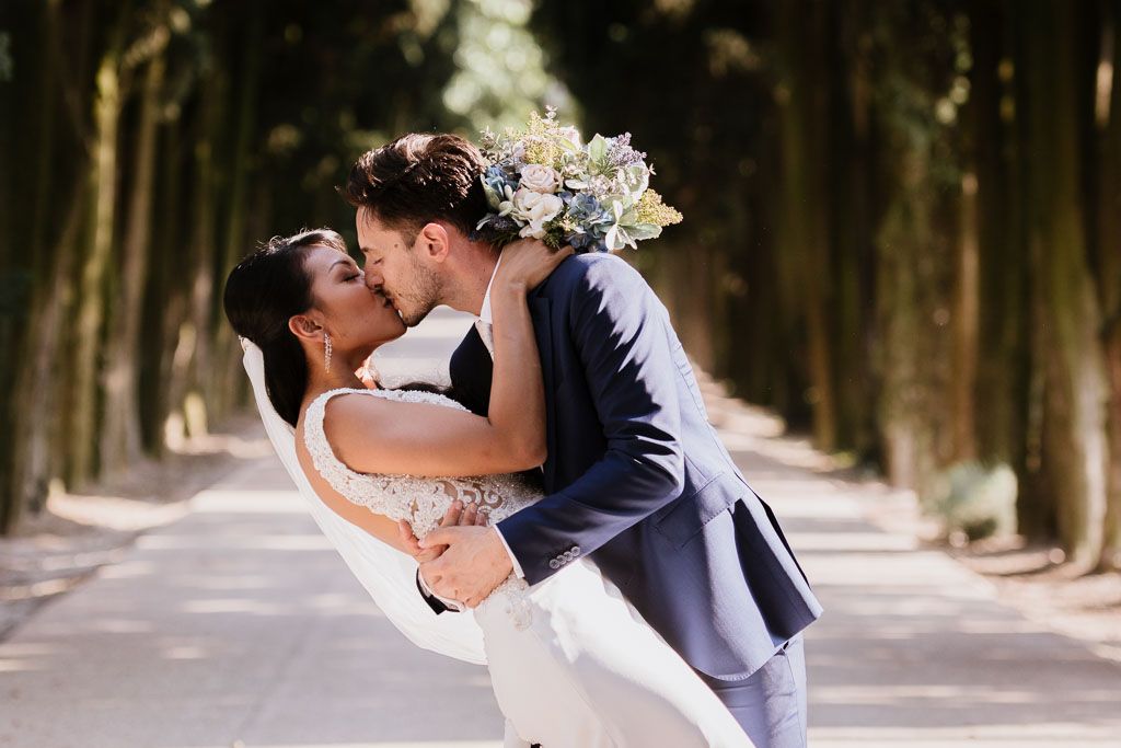 A romantic wedding couple dips while kissing on a tree-lined path beneath towering cypress trees.