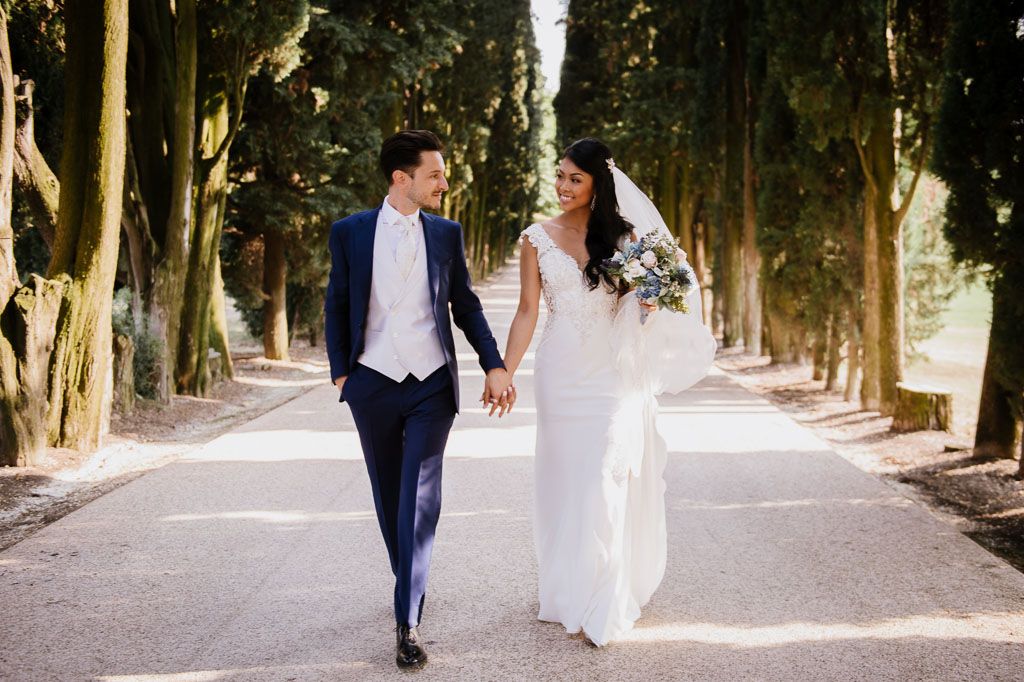 Newlyweds walk hand in hand down tree-lined path during outdoor wedding photoshoot.