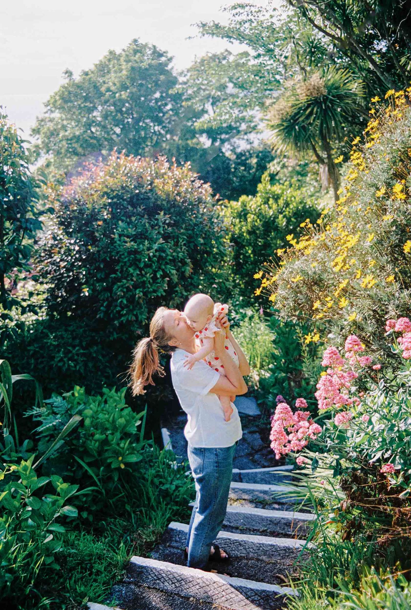 A couple embraces in a lush garden path with blooming pink flowers and green foliage on a sunny day.