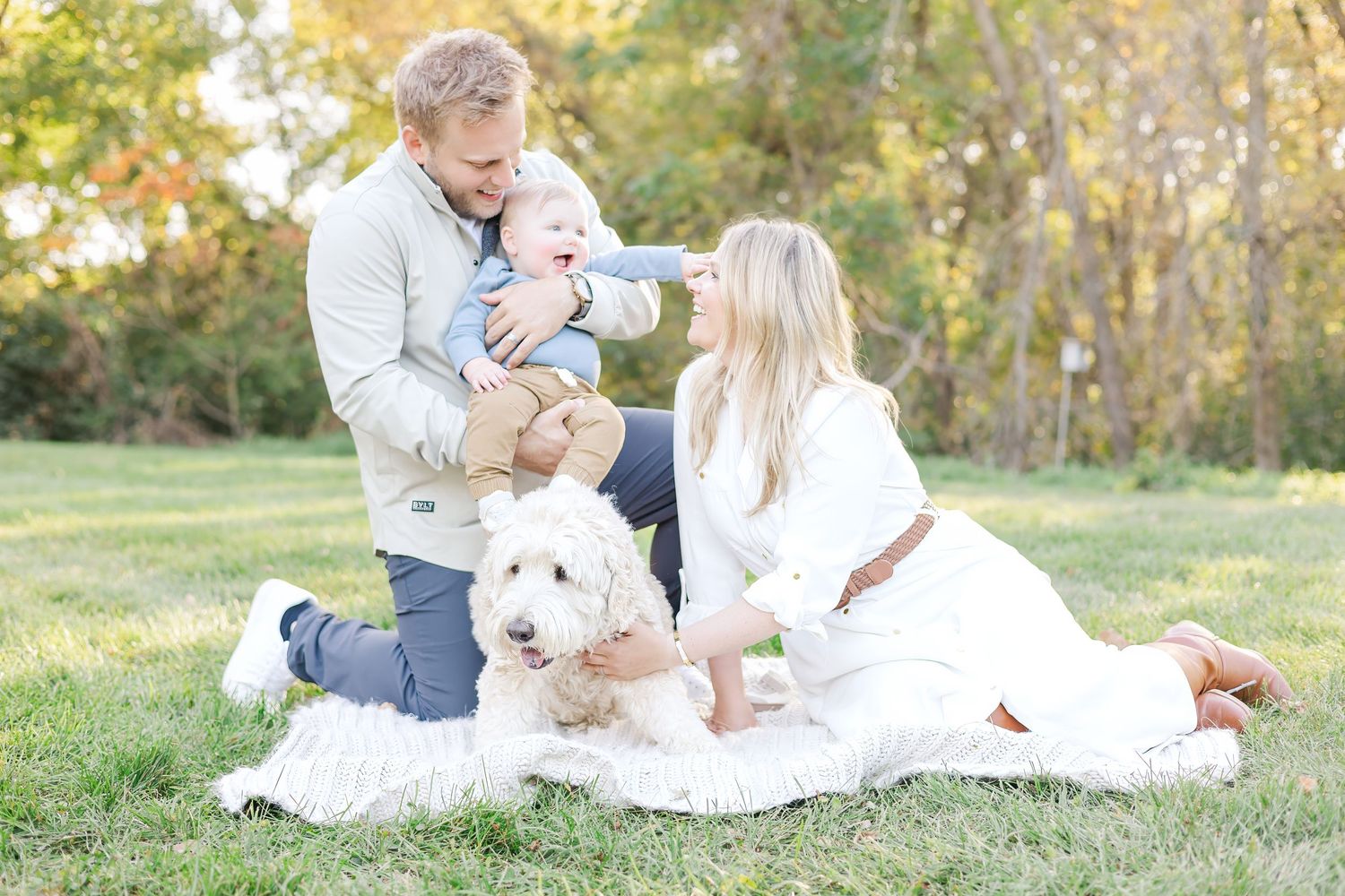 A couple sits on a blanket with their baby and white dog in an autumn field.
