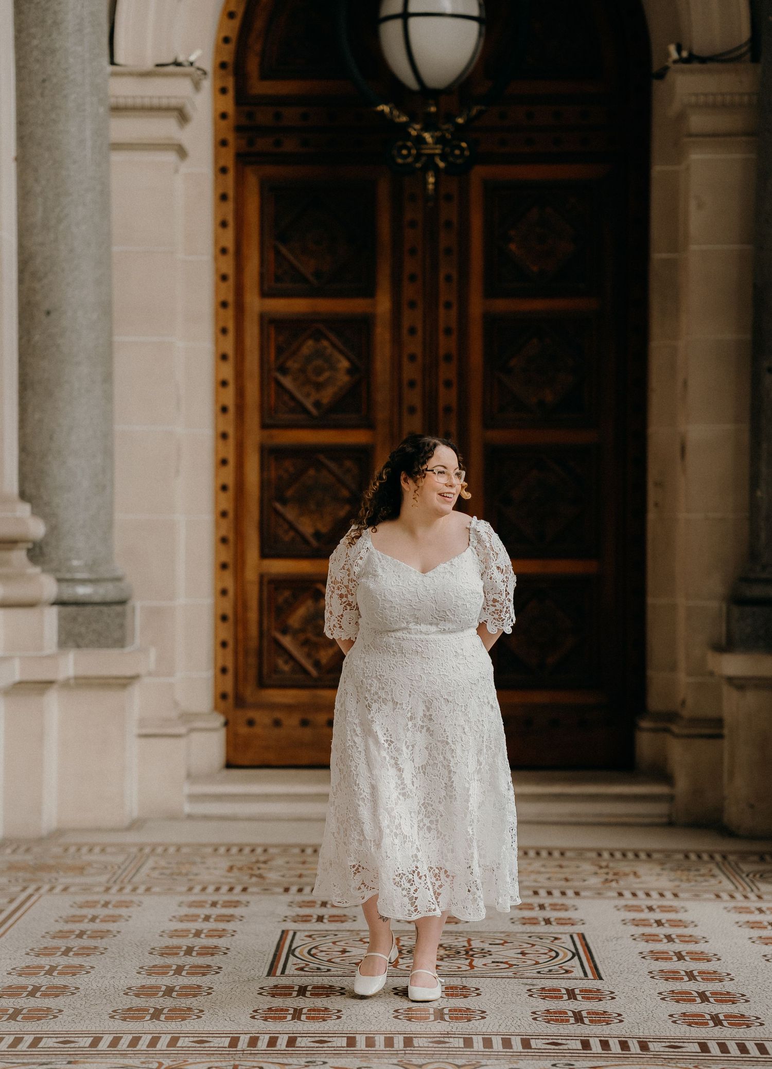 Elegant bride in white lace dress walks across ornate mosaic tile floor in historic building with stone columns.