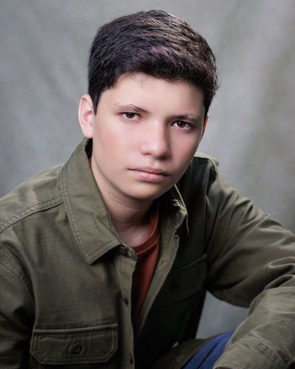 Studio portrait of a young person in an olive green military-style jacket against a gray background.