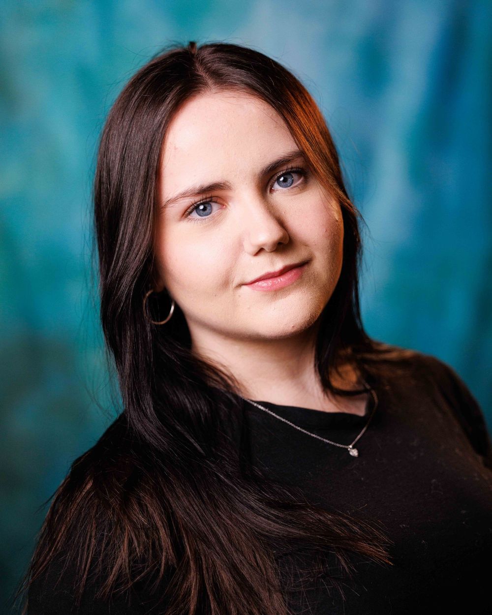 Young woman with long dark hair and blue eyes wearing a black top and necklace against a teal background.
