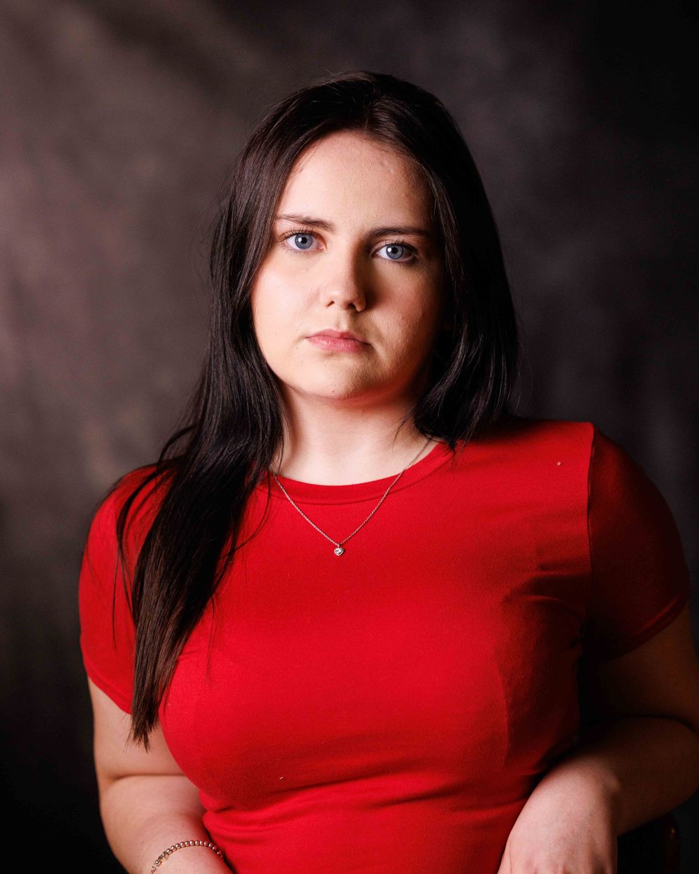 Young woman with dark hair and blue eyes wearing a red top and delicate necklace poses for a portrait.